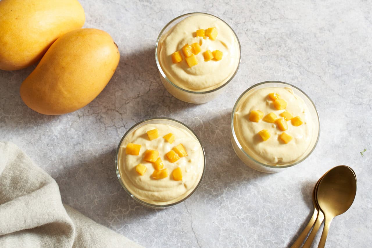 Three servings of mango mousse in small glasses topped with diced fresh mango, three gold spoons are on the right, two yellow mangoes are on the left.