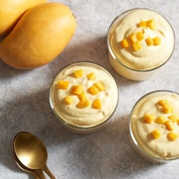 Three servings of mango mousse in small glasses topped with diced fresh mango, three gold spoons are on the right, two yellow mangos are on the left.