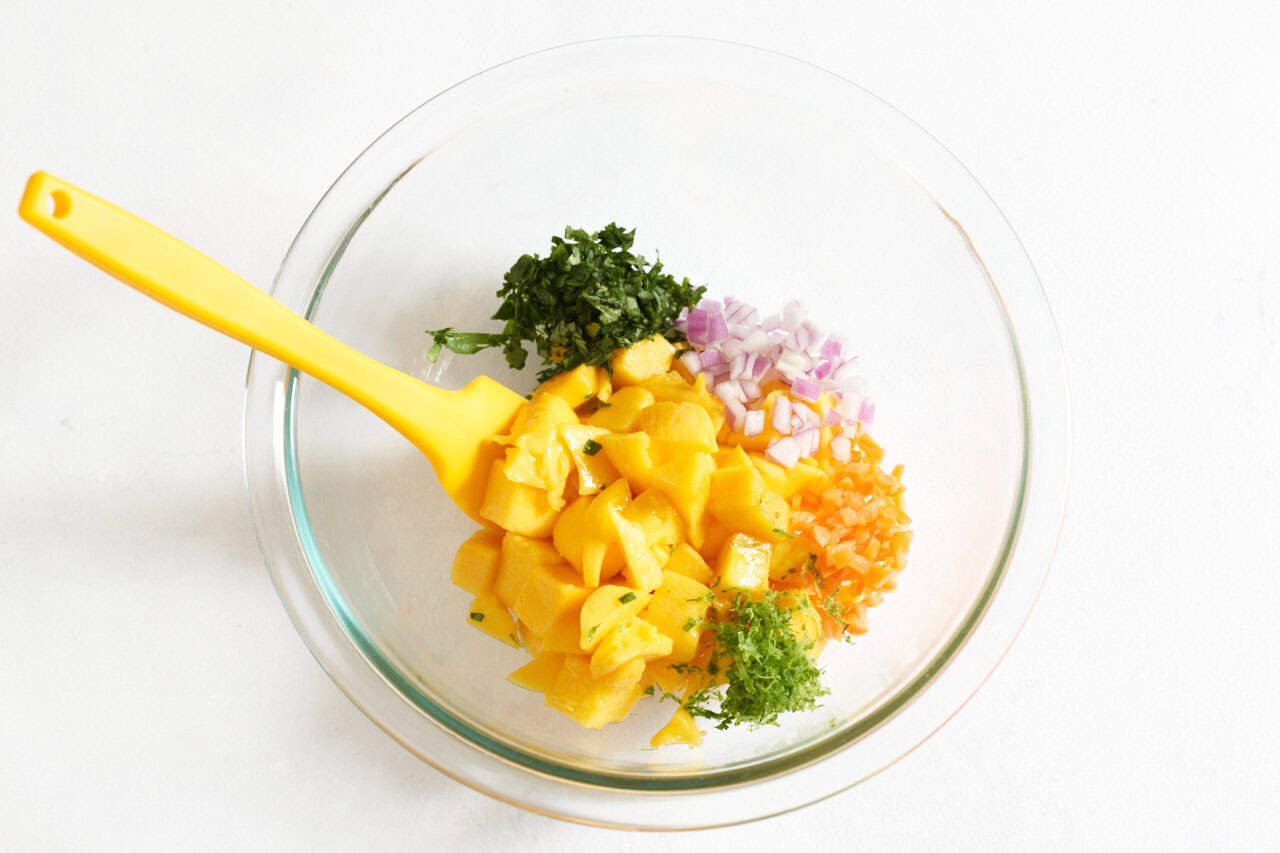 A yellow spatula in a bowl of fresh mango chunks with finely diced red onion, habanero pepper, lime zest, and cilantro.
