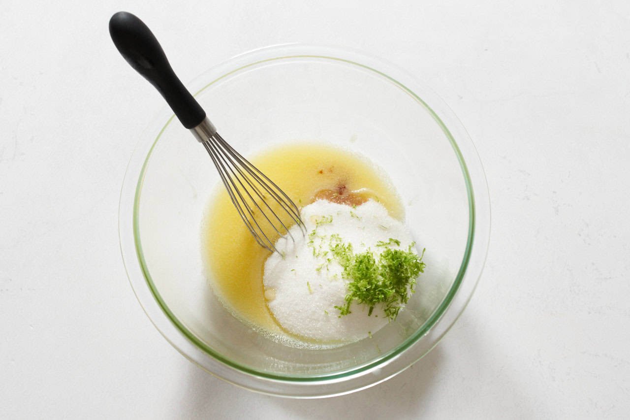 A whisk in a bowl of melted butter with sugar, vanilla and lime zest.