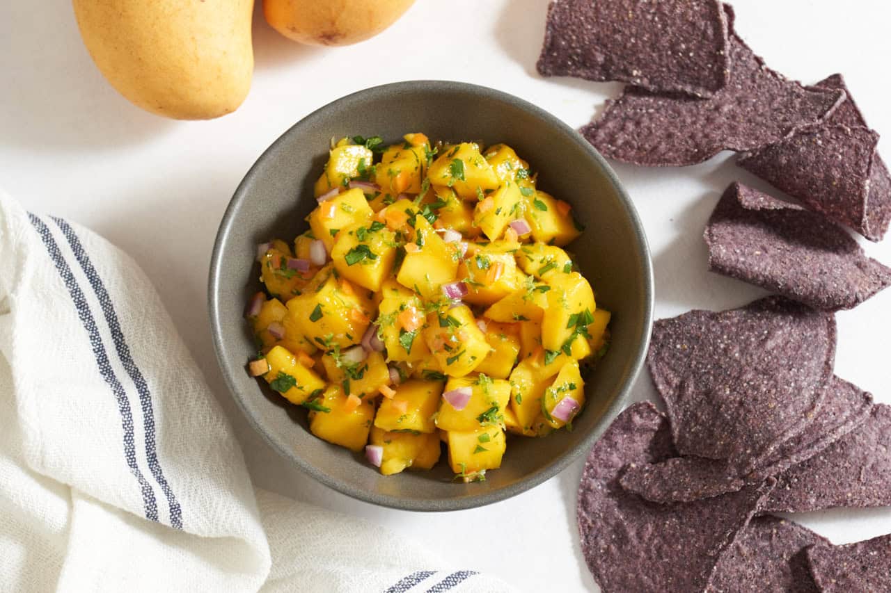A bowl of mango habanero salsa surrounded by blue corn tortilla chips, two mangoes, and a blue and white striped towel.