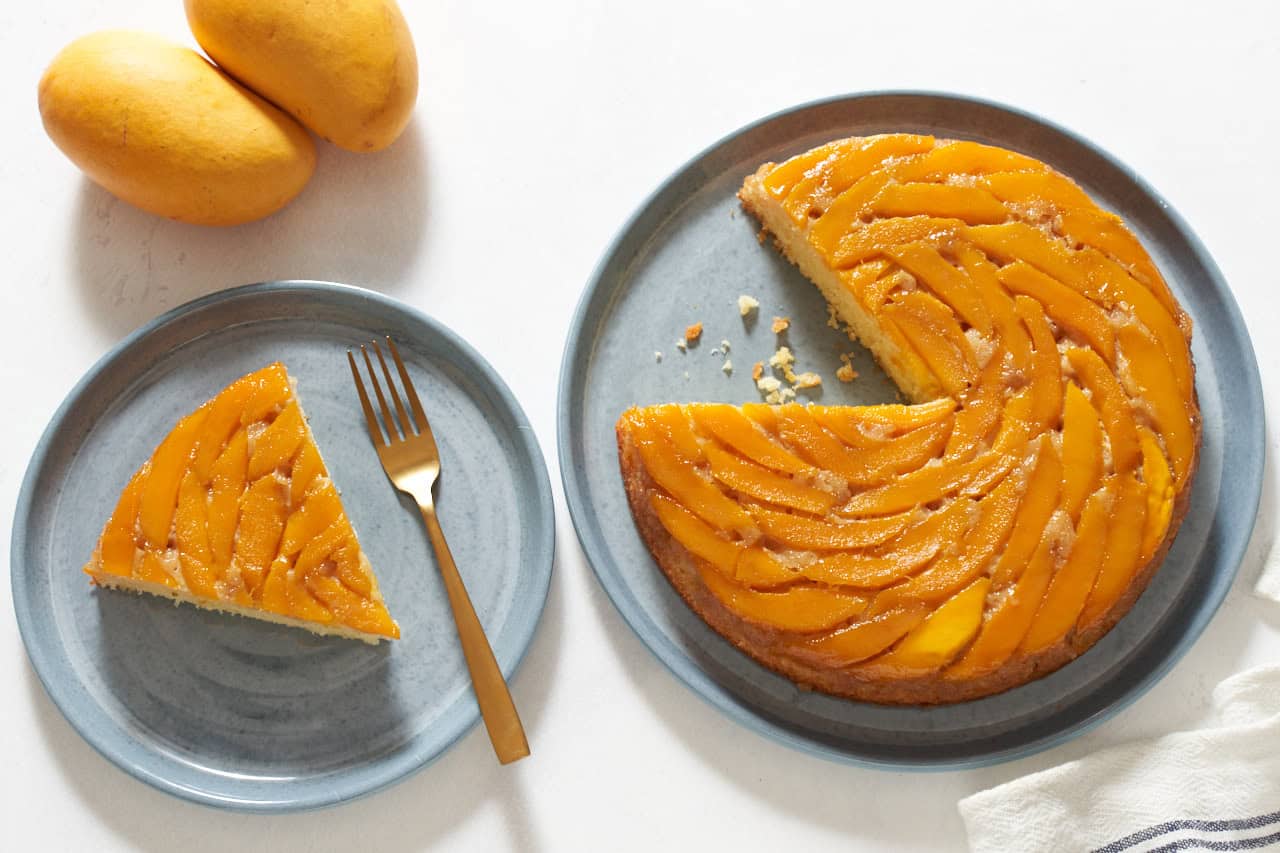 A mango upside down cake with a slice cut out of it on a blue plate, a plate with a slice of cake is on the left, two mangoes are in the upper left.
