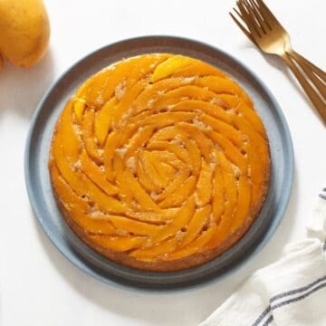 A mango upside down cake on a blue plate, gold forks are on the right, two mangoes are in the upper left.