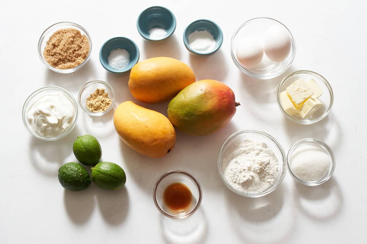 Three mangoes, three limes, and small bowls of flour, sugar, butter, eggs, ginger, brown sugar, yogurt, vanilla, salt, baking powder, and baking soda.