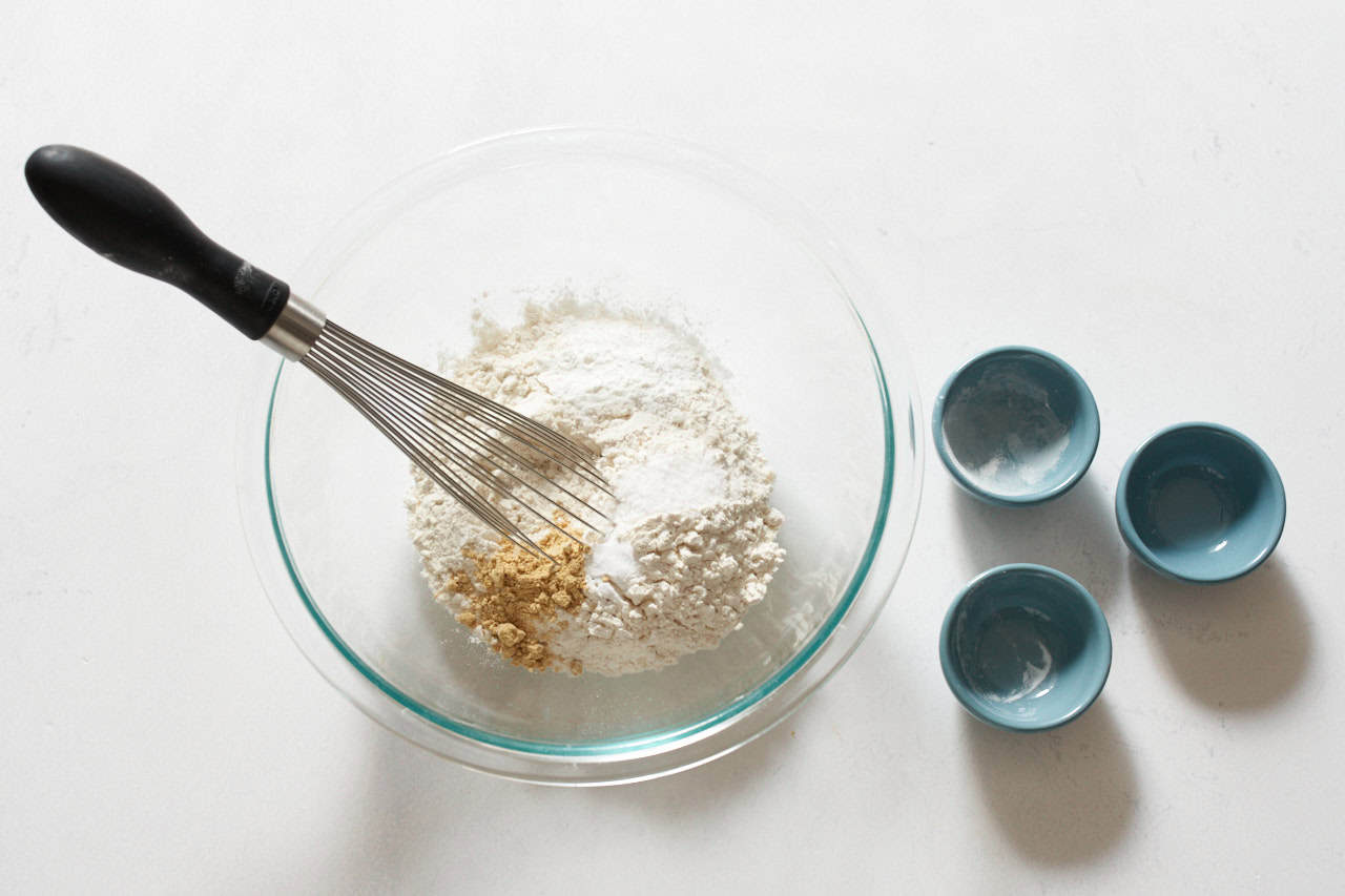 A whisk in a bowl of flour and other dry ingredients, three small blue bowls are on the right.