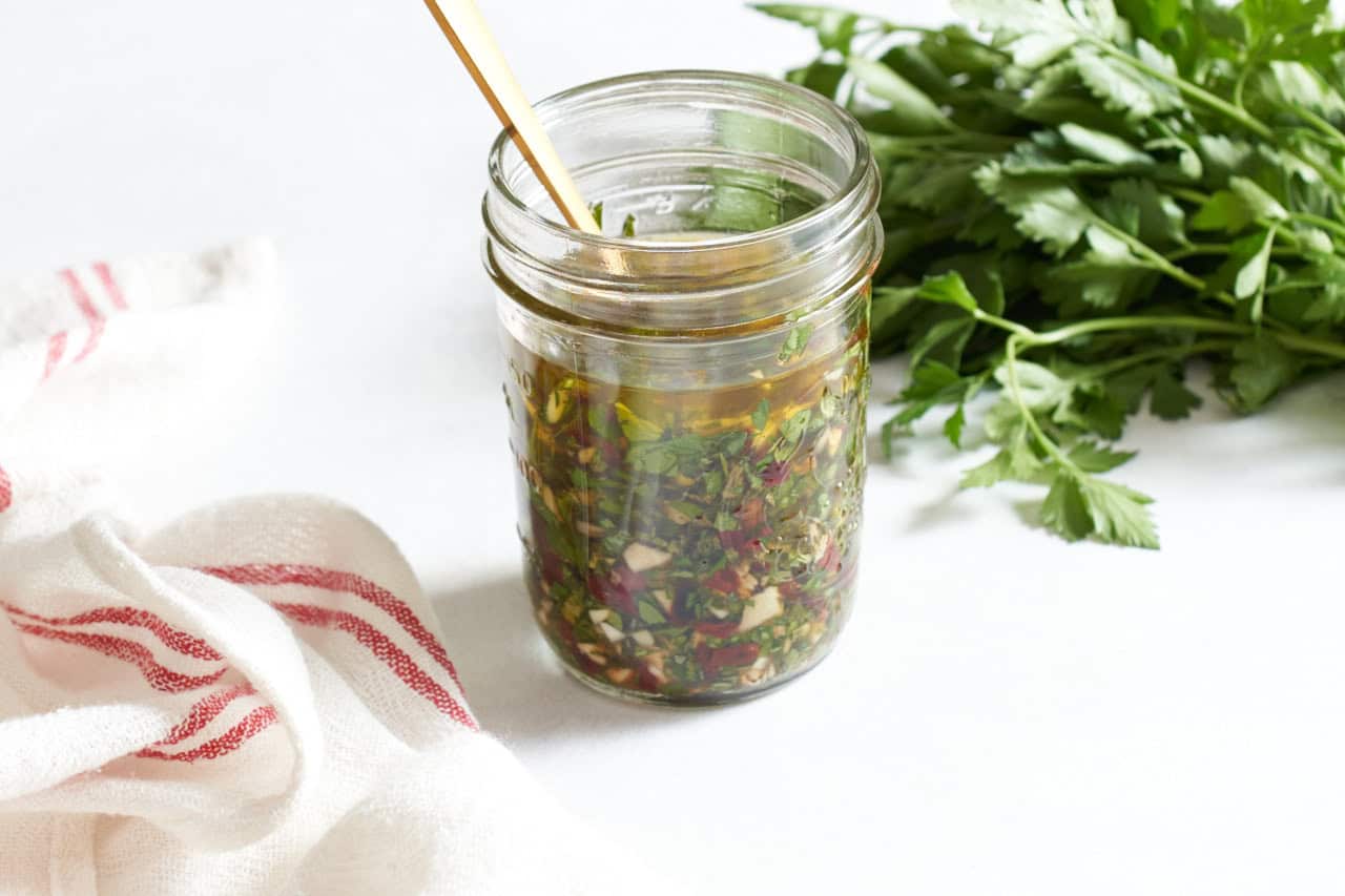 Argentine chimichurri sauce in a jar with a gold spoon in it, a bunch of fresh parsley is in the background.