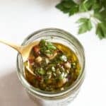 A spoonful of chimichurri sauce over a jar of the sauce, fresh parsley is in the upper right corner.