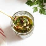 A spoonful of chimichurri sauce over a jar of the sauce, fresh parsley is in the upper right corner.