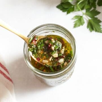 A spoonful of chimichurri sauce over a jar of the sauce, fresh parsley is in the upper right corner.