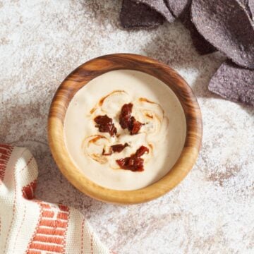 Chipotle crema topped with chopped chipotles in a wooden bowl, blue corn chips are in the upper right.