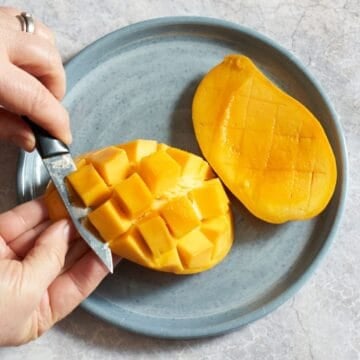 A woman's hands are shown slicing cube-shaped pieces of mango away from its peel, another mango half is on a plate to the right.