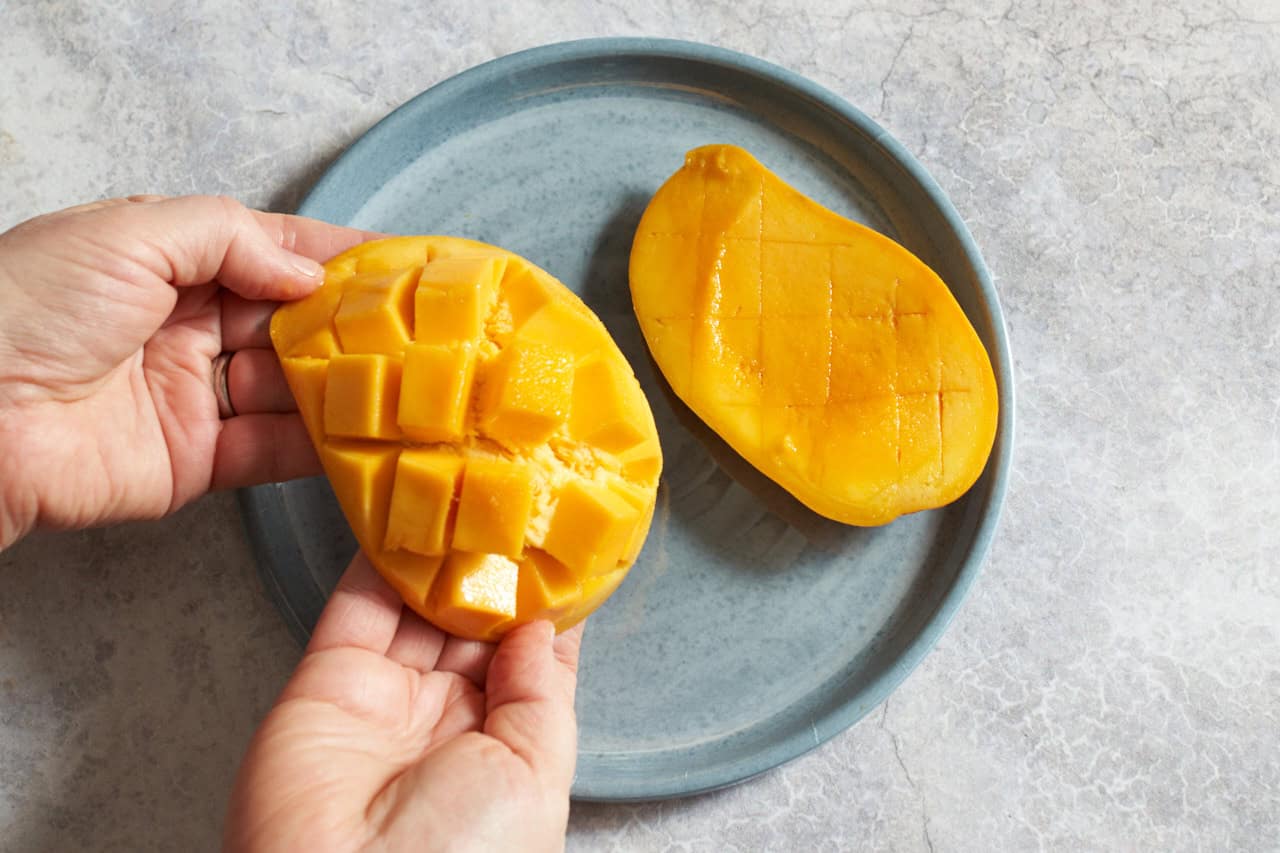 A woman's hands are shown bending a mango that has been scored so that cube-shaped pieces of the fruit are releasing from the peel, another mango half is on a plate to the right.