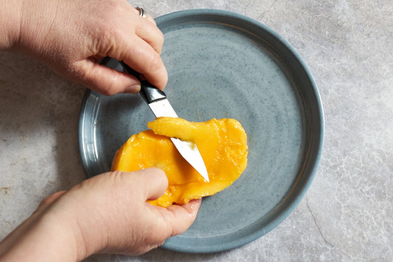 A woman's hands are shown using a paring knife to cut strips of fruit away from a mango seed.