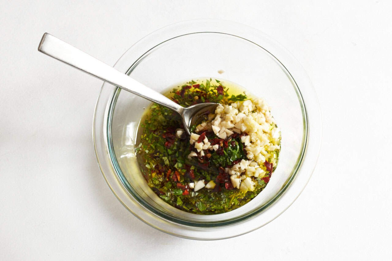 A mixing bowl of minced garlic, chopped parsley, and other ingredients for chimichurri sauce with a spoon in it ready to be mixed together.