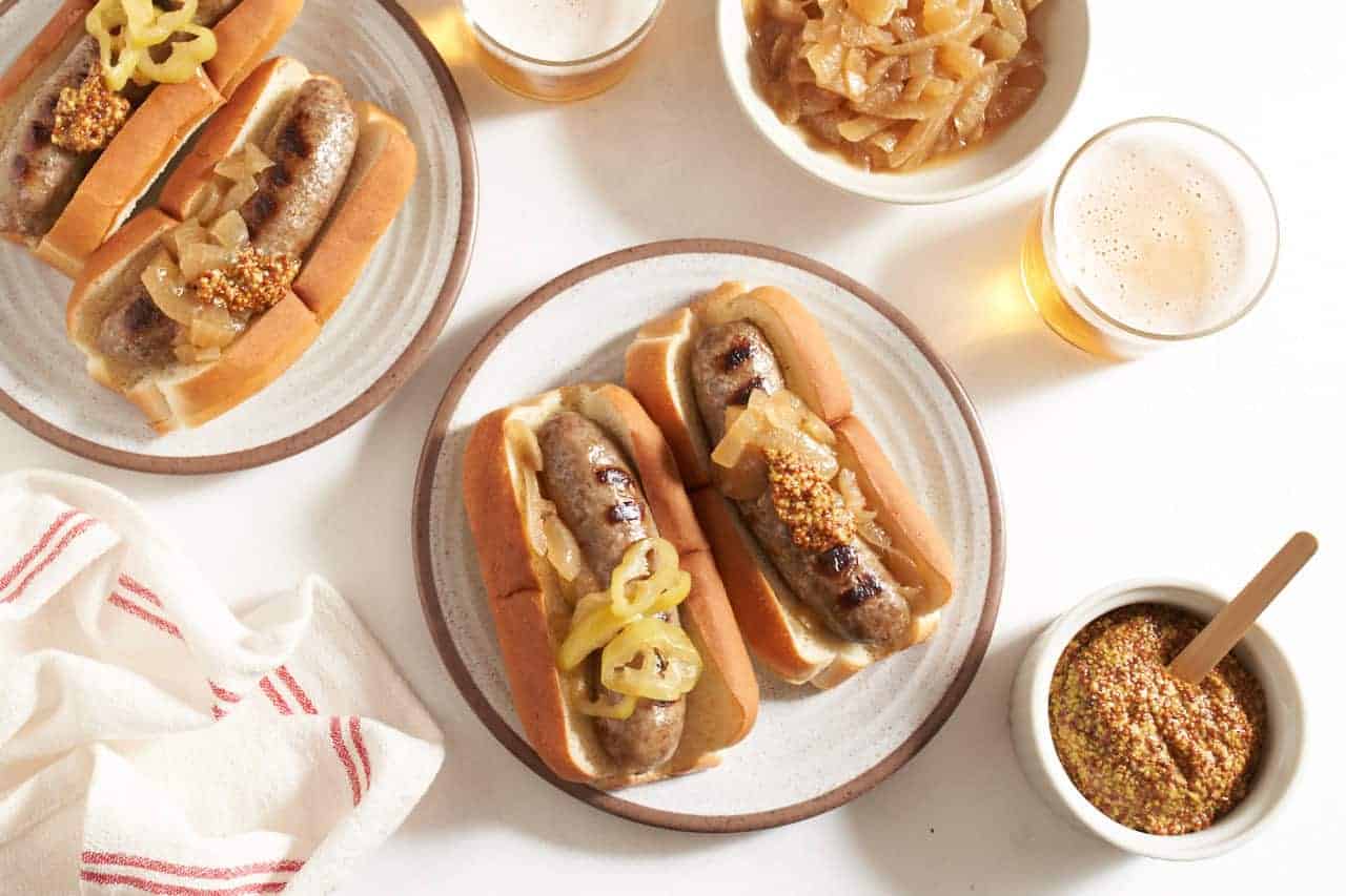 Two plates with beer brats in buns topped with onions, peppers, and mustard, two beers and small bowls of grainy mustard and onions are on the right.