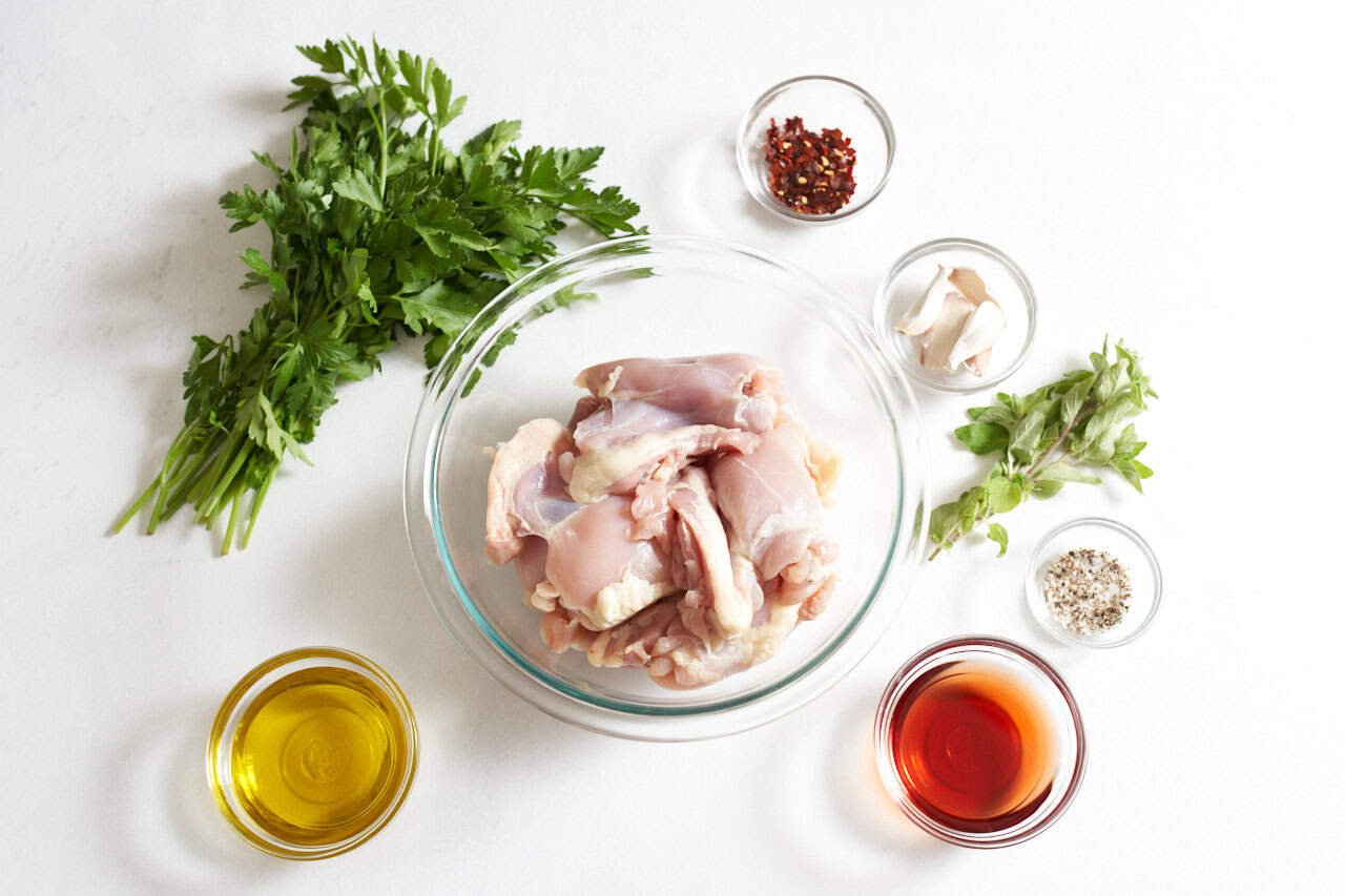 Fresh parsley and oregano, and bowls of chicken thighs, olive oil, red wine vinegar, garlic cloves, crushed red pepper, and salt and pepper.