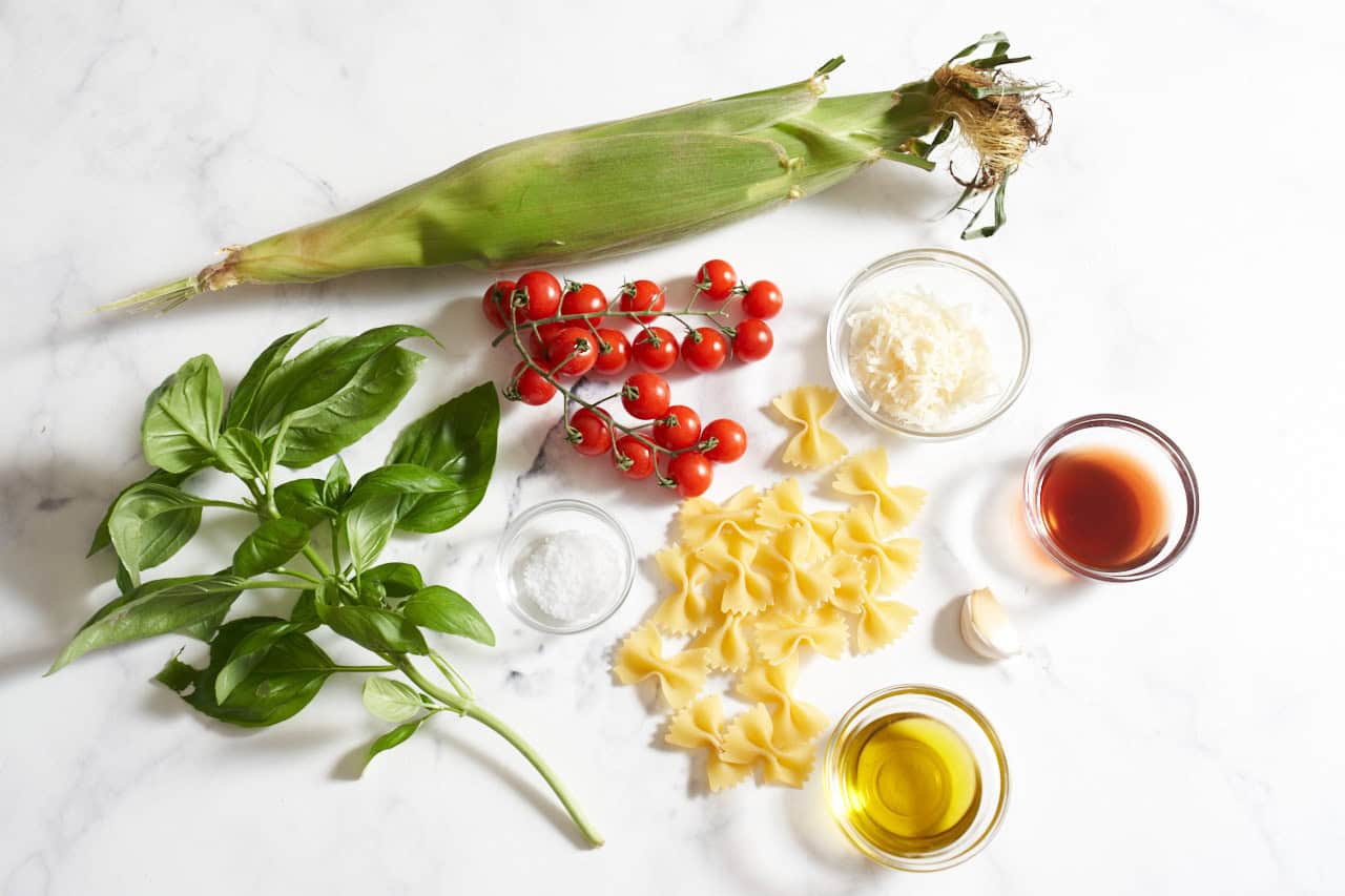 An ear of corn in the husk, fresh basil, cherry tomatoes on the vine, some bowtie pasta noodles, a clove of garlic, and small bowls of parmesan cheese, red wine vinegar, olive oil, and salt.