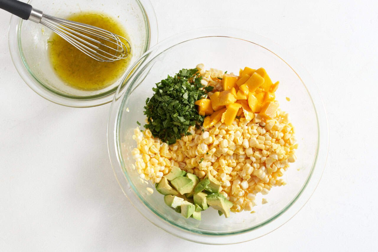 A bowl of fresh corn, diced mangoes and avocado and chopped cilantro next to a bowl of salad dressing with a whisk in it.