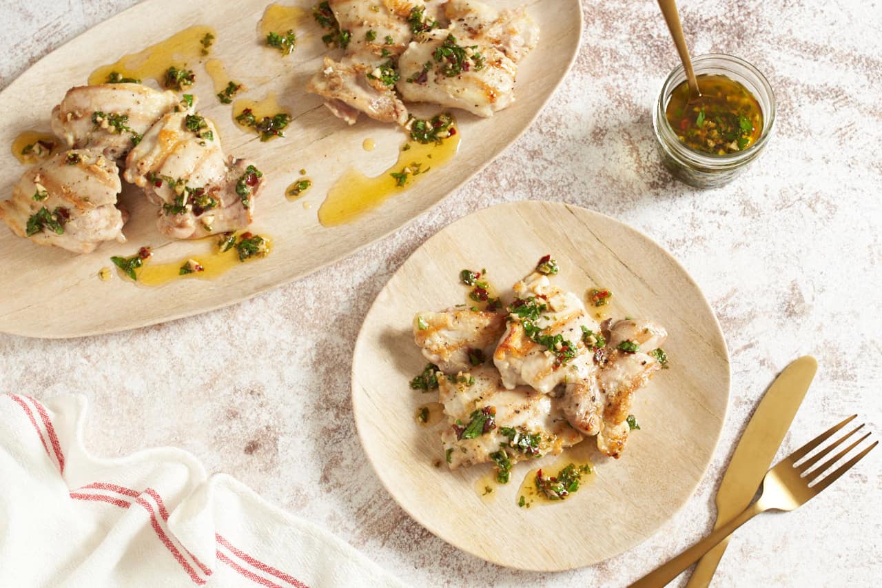 A small round plate of grilled chicken with chimichurri sauce next to an oval platter of the same, a small jar of chimichurri is to the right, gold utensils are in the bottom right corner.