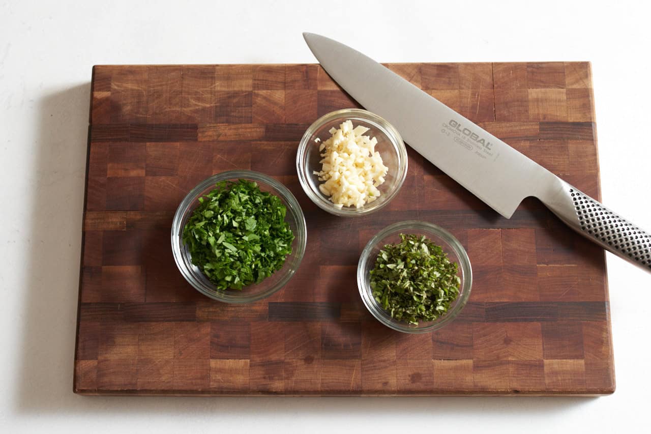 A knife on a cutting board with small bowls of finely chopped parsley, oregano, and garlic.