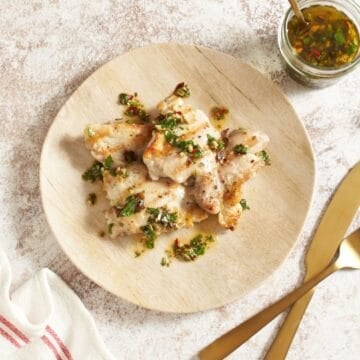 A small round plate of grilled chicken with chimichurri sauce, a small jar of chimichurri is to the right, gold utensils are in the bottom right corner.