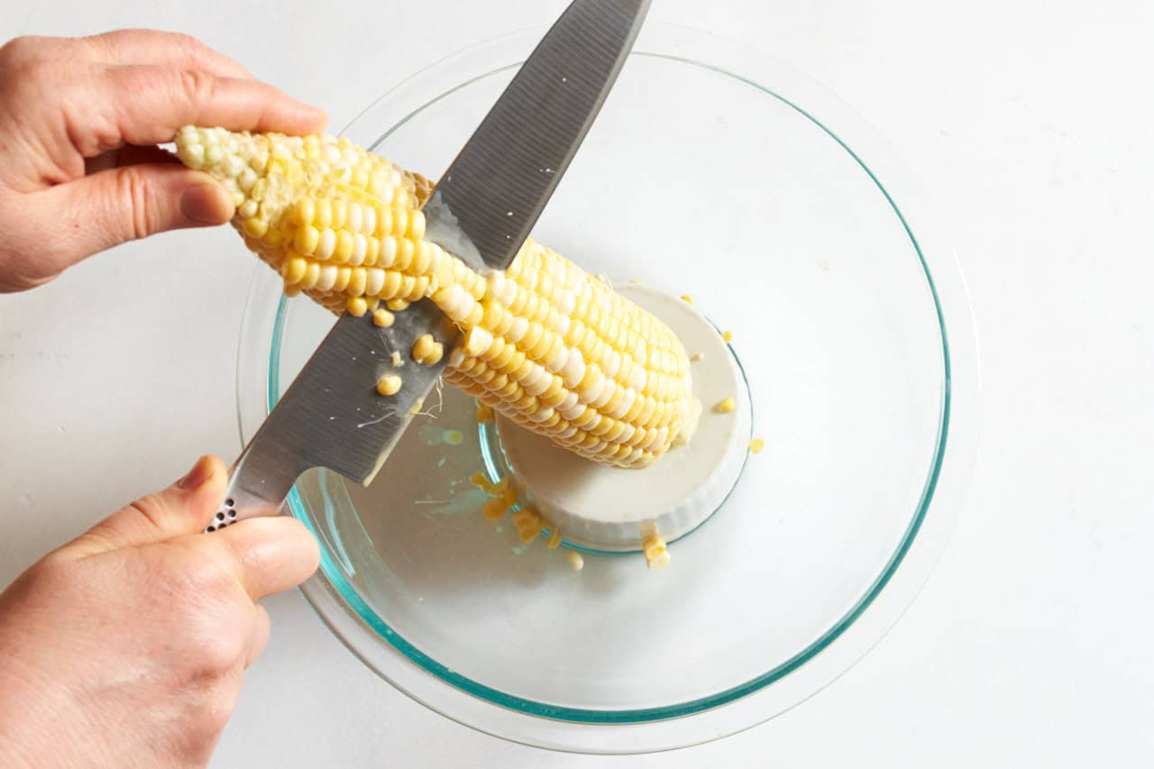 A woman's hands shown cutting corn off the cob with a chef's knife by standing it up on a small bowl set inside a larger one.