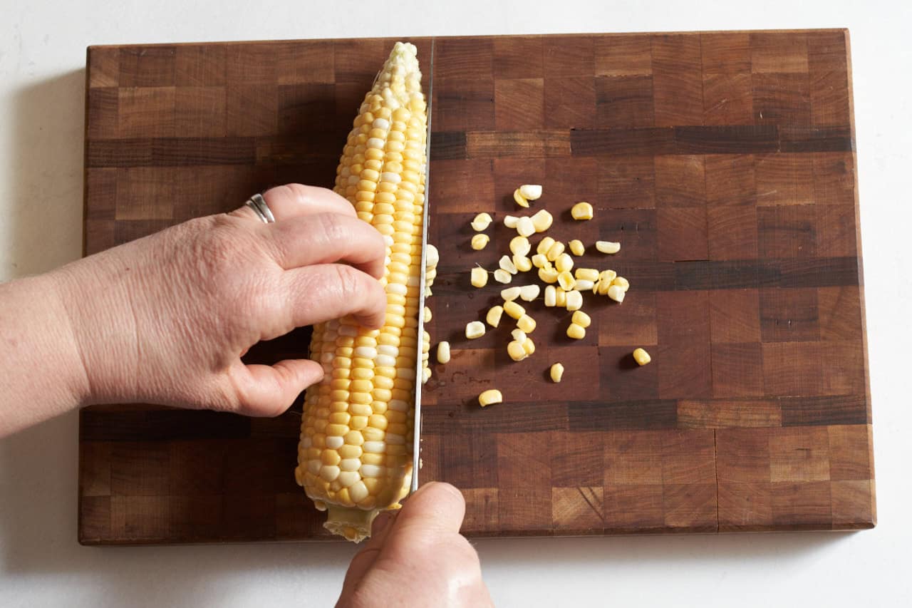 A woman's hands shown cutting corn off the cob with a chef's knife by laying it flat on a wooden cutting board.