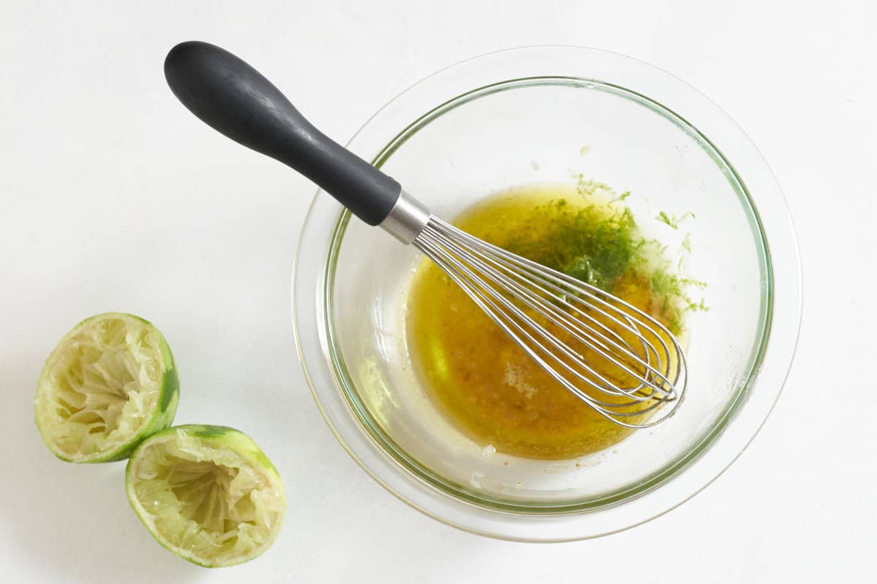 A whisk in a bowl of lime vinaigrette, two squeezed lime halves are on the right.