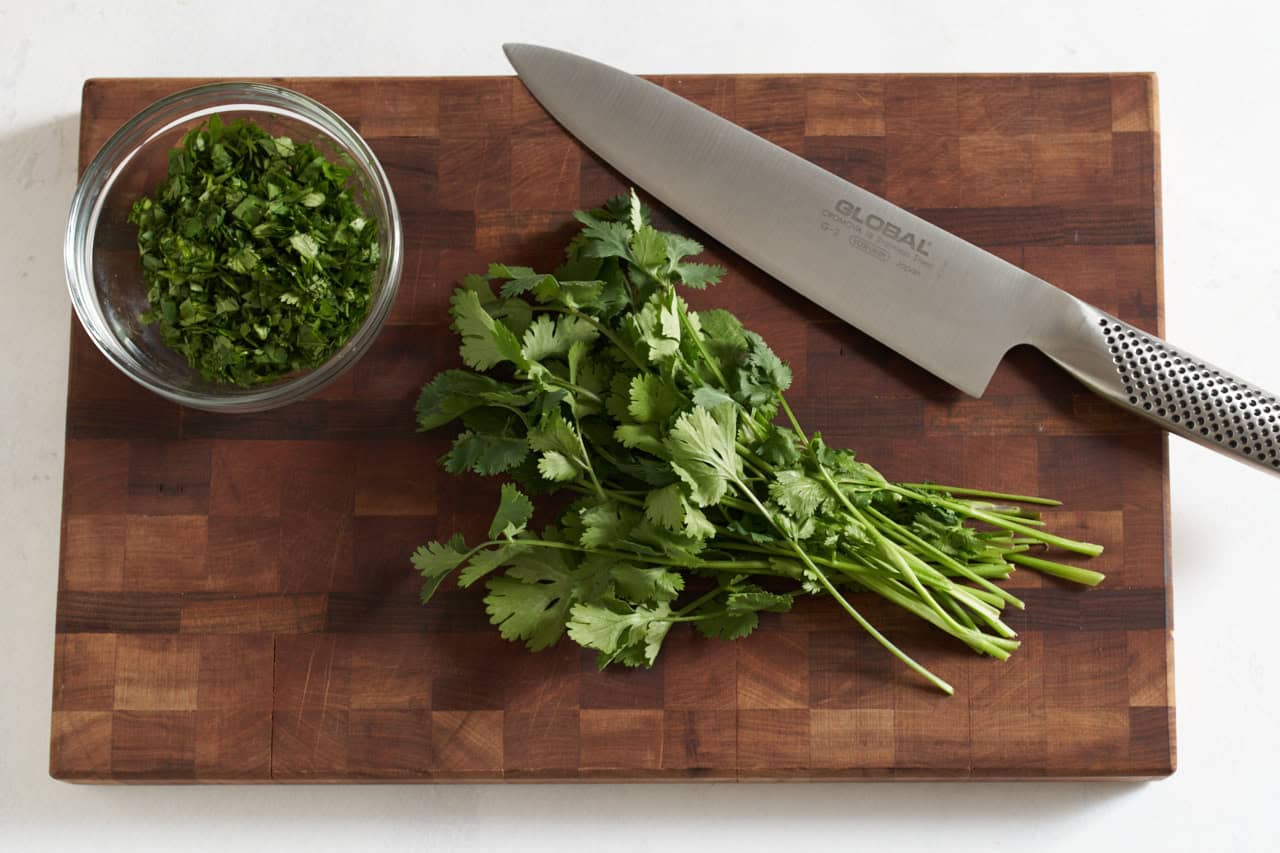 A knife on a cutting board with fresh cilantro and some chopped cilantro in a small glass bowl.