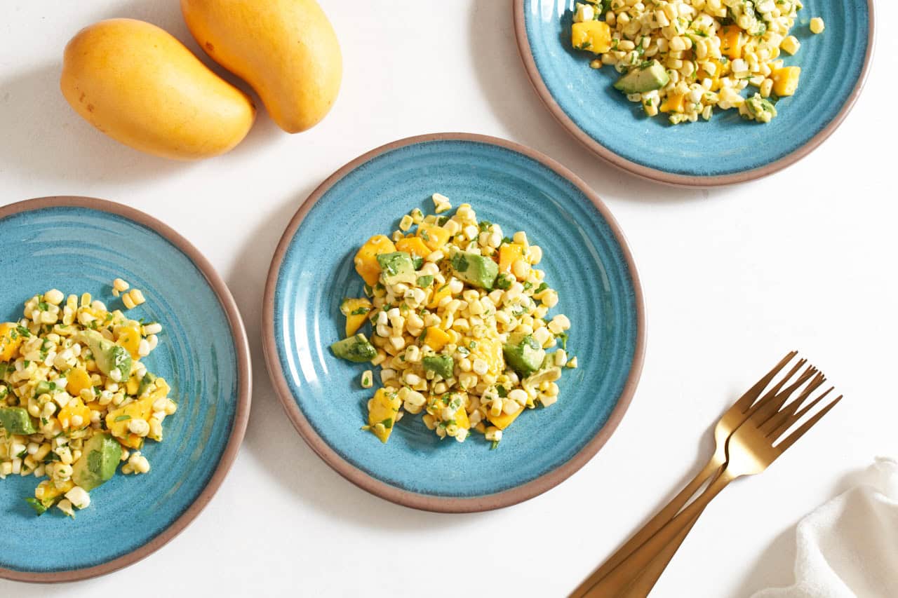 Three blue plates with mango corn salad on them, gold forks are on the right, two mangoes are in the upper left.