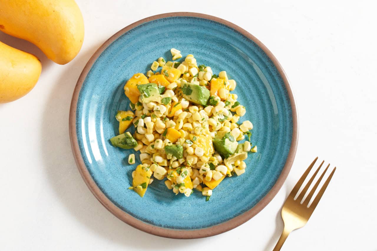 A blue plate with mango corn salad on it, a gold fork is on the right, two mangoes are in the upper left.