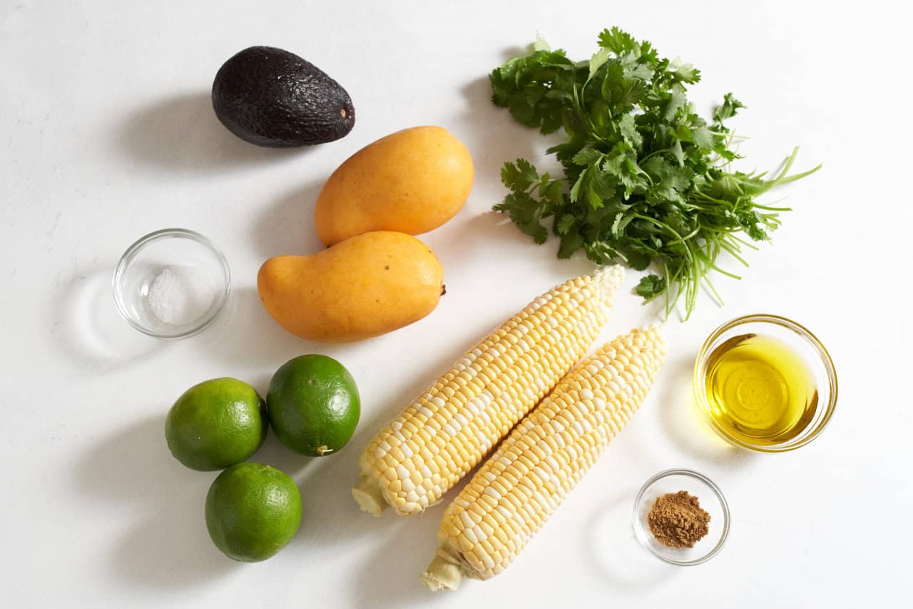 Two ears of shucked corn, two mangos, three limes, an avocado, a bunch of cilantro, and small bowls with olive oil, salt, and cumin.