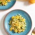 Two blue plates with mango corn salad on them, a gold fork is on the right, two mangoes are in the upper right.
