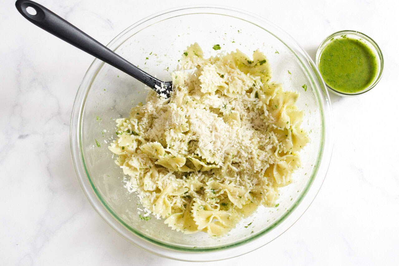 A spatula in a bowl of bowtie pasta topped with parmesan, a small bowl of basil dressing is on the right.