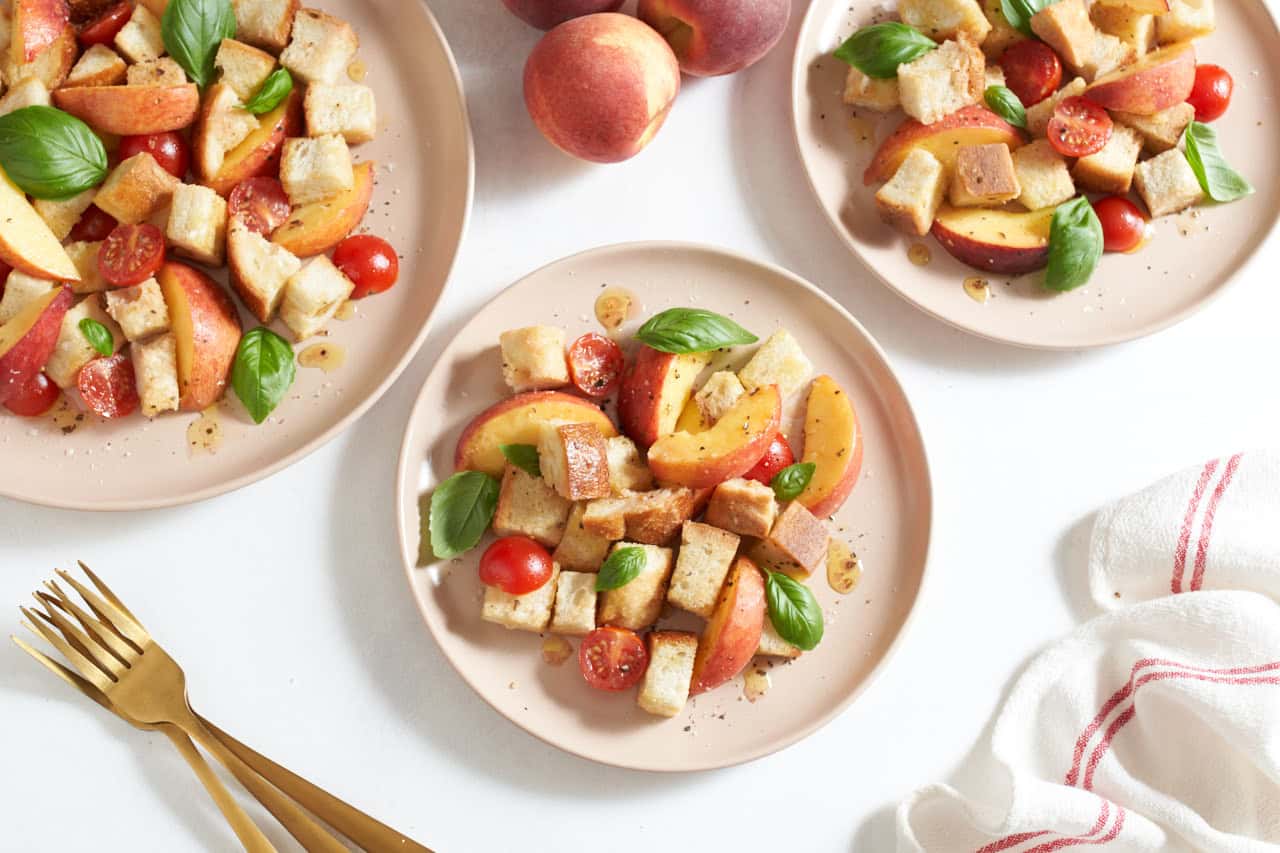 Three pink plates of peach panzanella salad topped with fresh basil leaves, gold forks are on the left, a red and white striped towel is on the right.