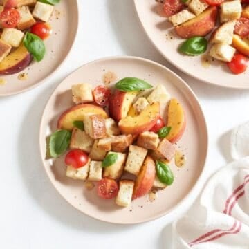 Three pink plates of peach panzanella salad topped with fresh basil leaves.