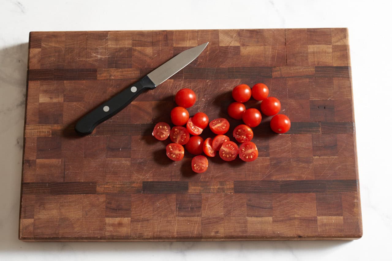 A paring knife on a wooden cutting board with cherry tomatoes that are sliced in half.