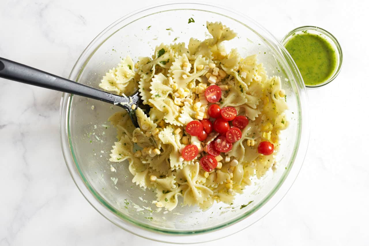 A spatula in a bowl of bowtie pasta with corn and cherry tomatoes mixed into it, a small bowl of basil dressing is on the right.