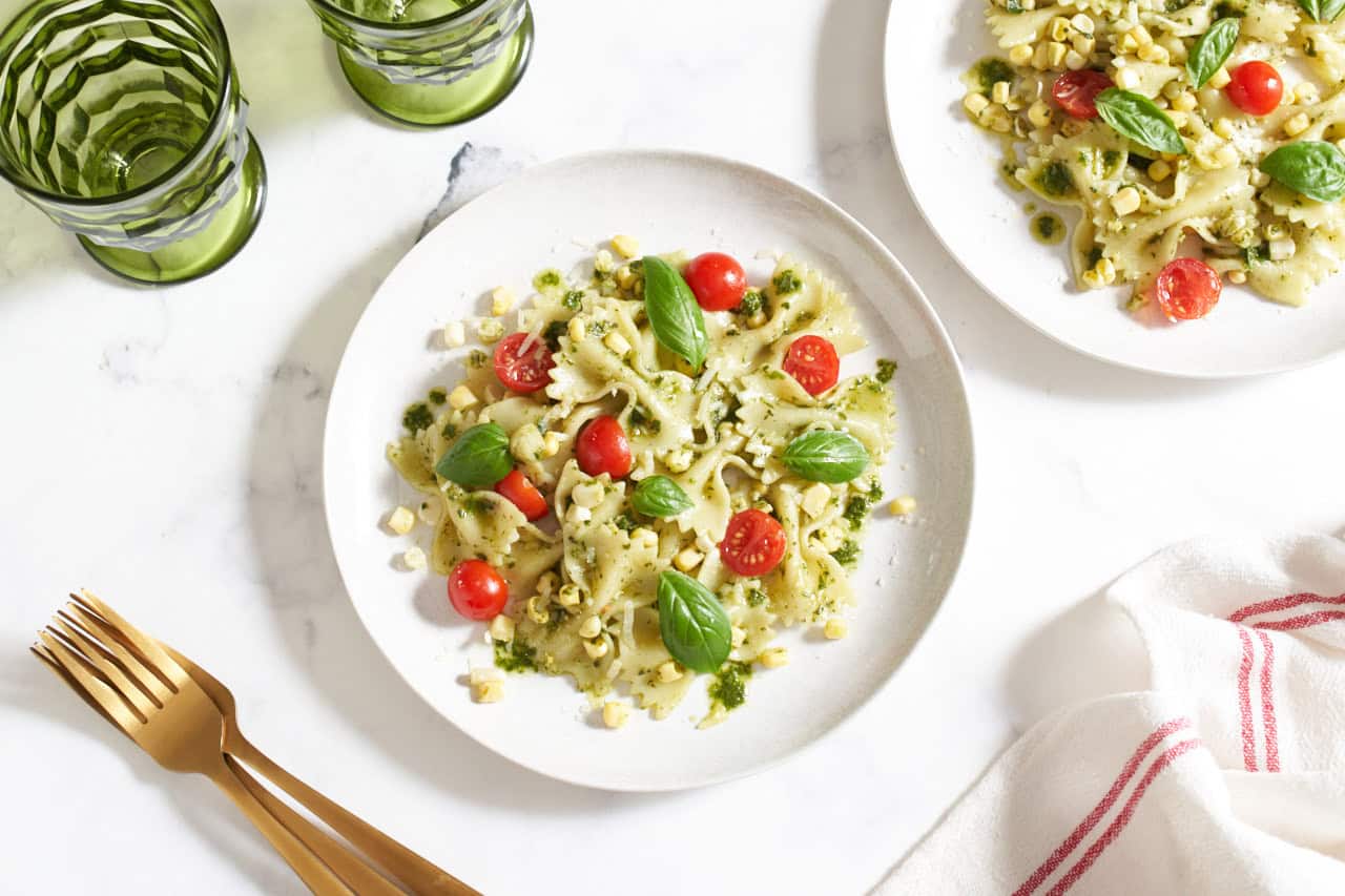 Two plates of summer corn pasta salad topped with cherry tomatoes and fresh basil, a red and white striped towel is on the right, gold forks and green glasses are on the left.