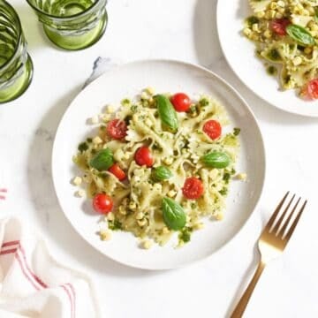 Two plates of summer corn pasta salad topped with cherry tomatoes and fresh basil, a red and white striped towel is on the right, gold forks and green glasses are on the left.