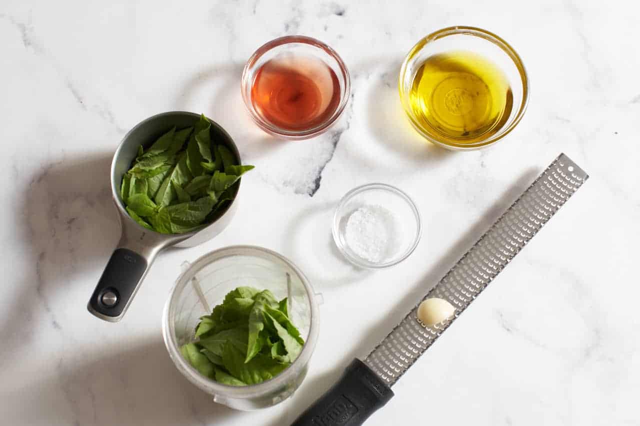 A small measuring cup and blender full of fresh basil leaves, three small bowls of olive oil, red wine vinegar and salt, and a microplane grater with a garlic clove on it.