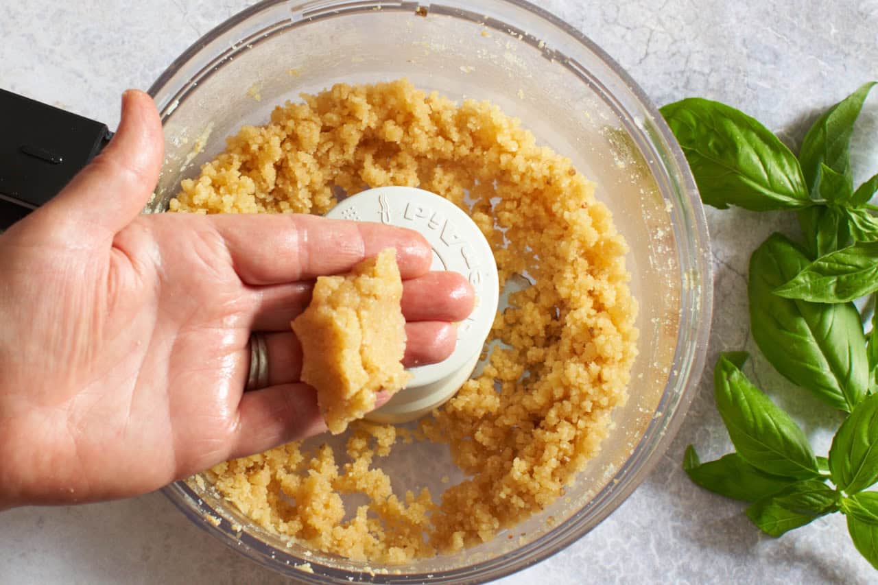 A woman's hand over a food processor holding a mixture of pine nuts and parmesan cheese.