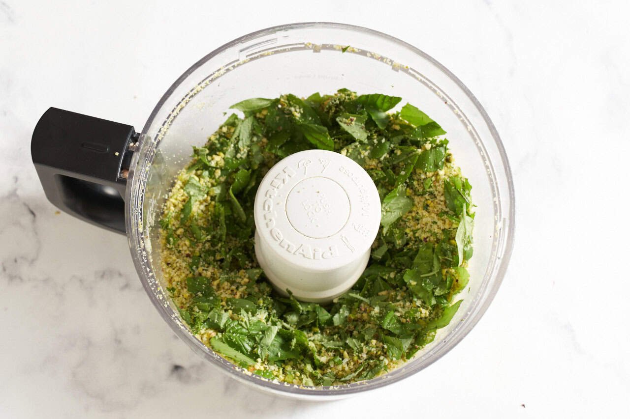 A food processor with pistachios, parmesan, and fresh basil that has been pulsed.