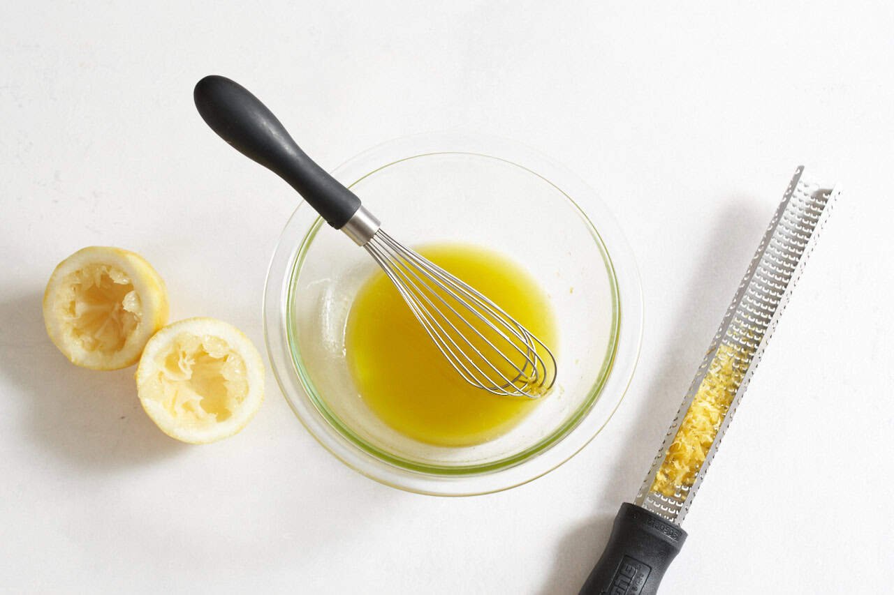 A whisk in a bowl of lemon vinaigrette dressing, a microplane grater with lemon zest and two lemon halves are on either side.