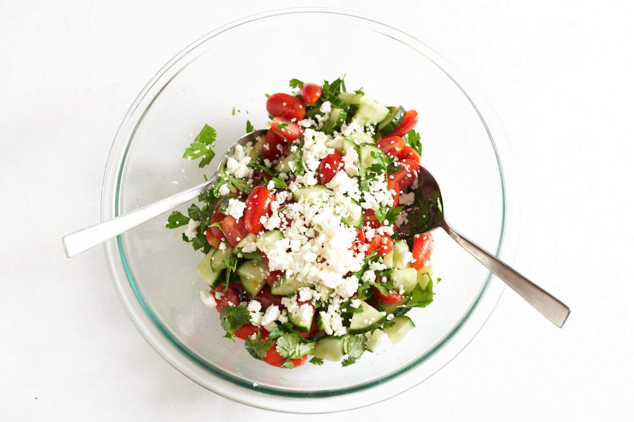 Cucumber tomato salad topped with feta cheese, two spoons are on each side of the bowl to be used for tossing the salad.