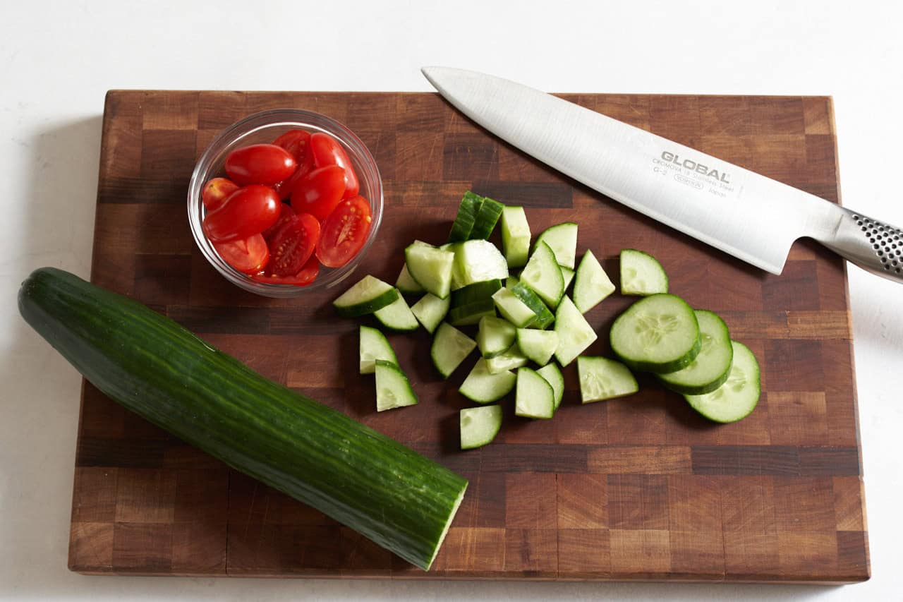 A knife on a wooden cutting board with a small bowl of cherry tomatoes cut in half, and cucumber slices that have been cut into quarters.