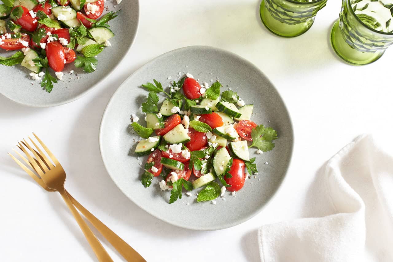 Two plates of cucumber, tomato, and feta salad, two gold forks are on the left, two green glasses are on the right.