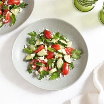 Two plates of cucumber, tomato, and feta salad, two green glasses are in the upper right.