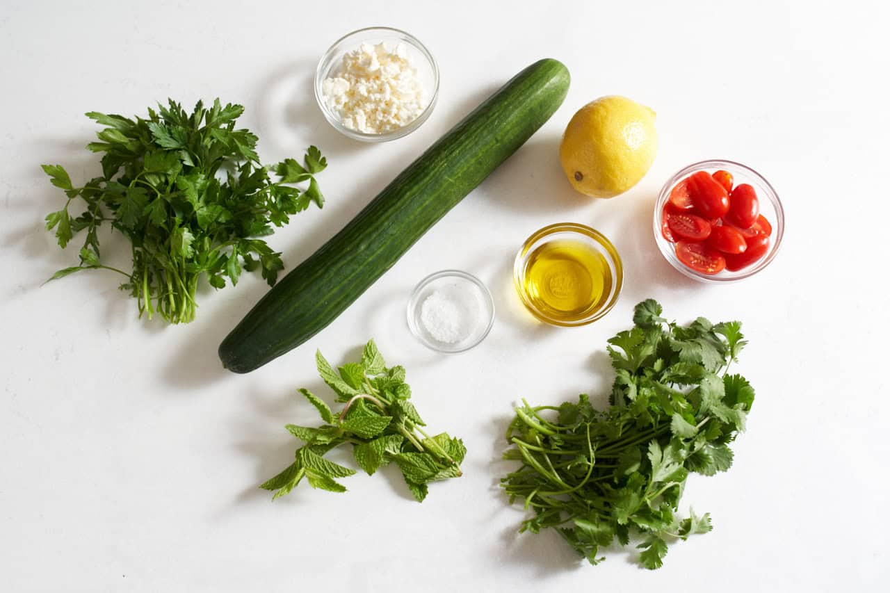 A cucumber, one lemon, fresh parsley, mint, and cilantro, three small glass bowls of sliced tomatoes, feta cheese, olive oil, and kosher salt.