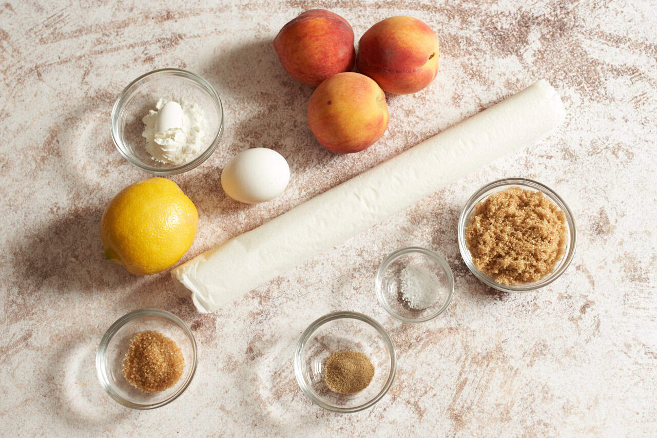Three peaches, a lemon, one egg, and small bowls of cardamom, brown sugar, salt, corn starch and turbinado sugar.